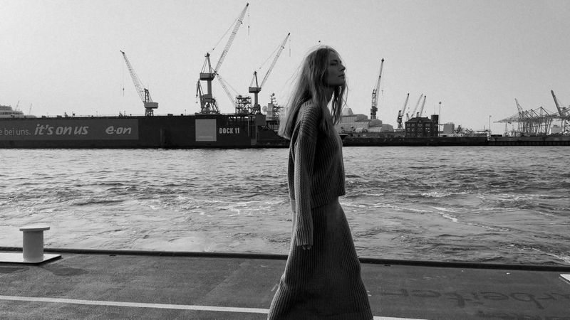Black and white fashion shot: a woman in a knit outfit walks along a pier, with cranes and ships visible in the background.