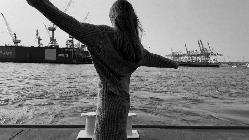 Black and white shot of a woman in knitwear with arms outstretched looking at cranes and cargo ships in a harbor.