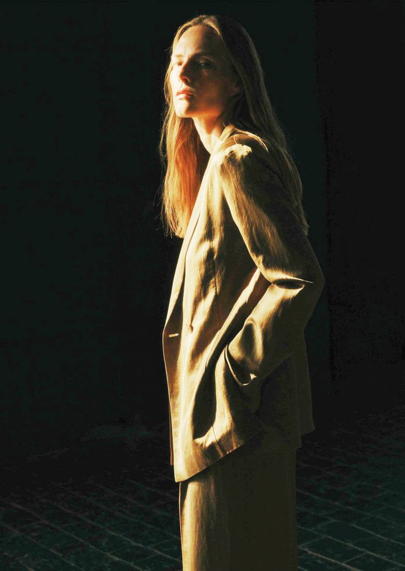 Woman in a tan blazer and pants, illuminated by a beam of sunlight against a dark background, looking slightly away from the camera.