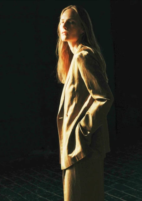 Woman in a tan blazer and pants, illuminated by a beam of sunlight against a dark background, looking slightly away from the camera.
