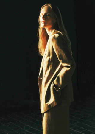 Woman in a tan blazer and pants, illuminated by a beam of sunlight against a dark background, looking slightly away from the camera.