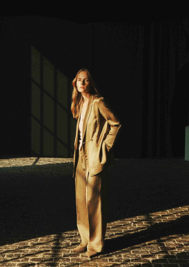 A woman in a brown suit stands in a sunlit room with a brick floor and a dark background, looking at the camera with a neutral expression.