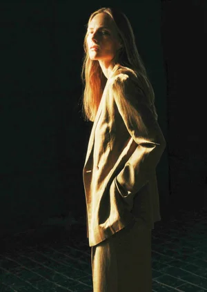 Model standing in profile wearing a neutral-toned blazer with warm sunlight illuminating her face against a dark background.