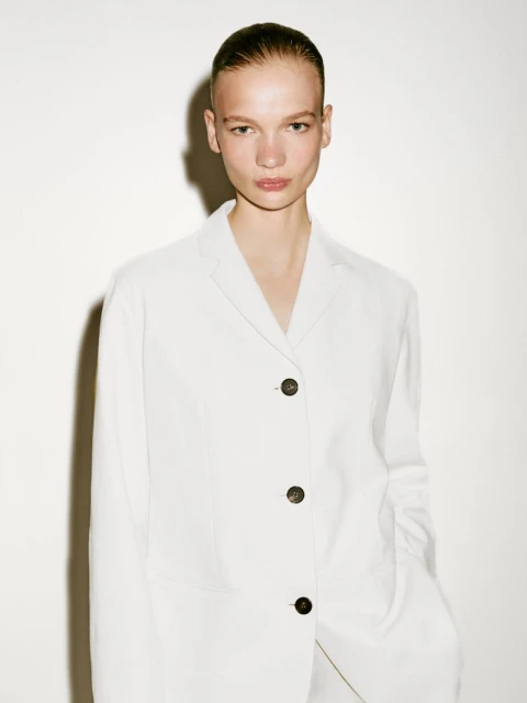 Close-up of an adult female fashion model in a white linen jacket looking directly at the camera against a white background.