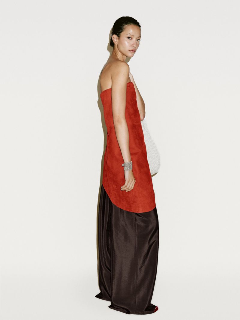 A slender woman poses in a fashion shot, wearing a strapless two-tone dress, with an orange bodice and a dark brown skirt, set against a white backdrop.