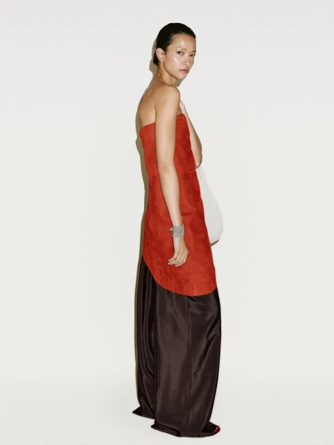A slender woman poses in a fashion shot, wearing a strapless two-tone dress, with an orange bodice and a dark brown skirt, set against a white backdrop.