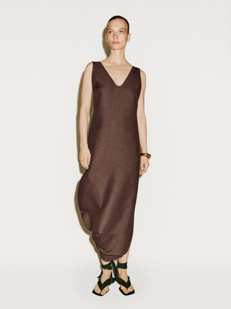 Elegant woman in a brown V-neck dress and stylish strappy sandals poses against a pristine white backdrop, showcasing a minimalist fashion aesthetic.