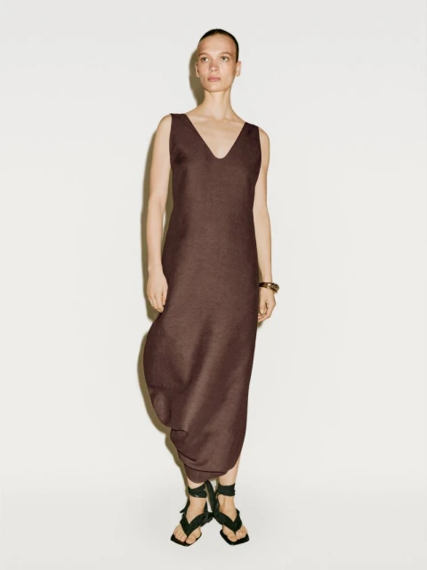 Elegant woman in a brown V-neck dress and stylish strappy sandals poses against a pristine white backdrop, showcasing a minimalist fashion aesthetic.