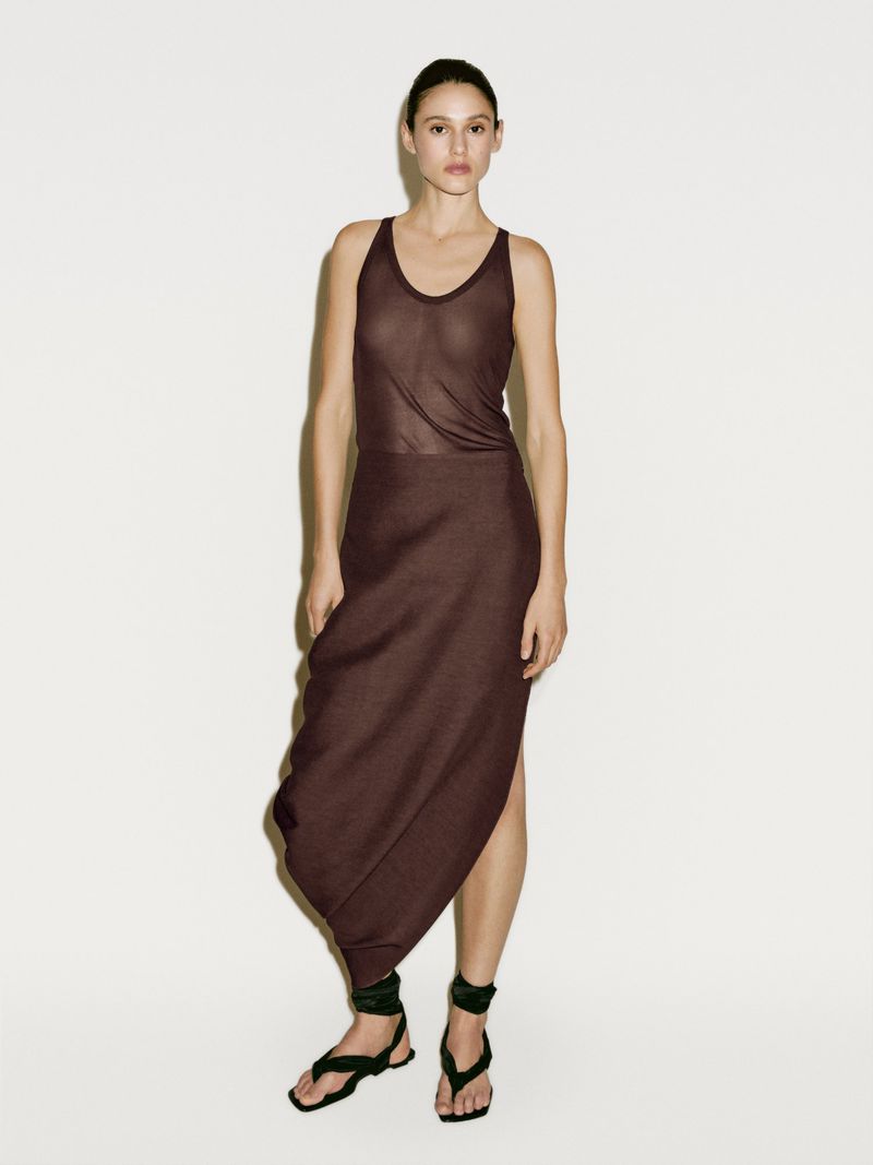 A woman in a dark brown tank top and a skirt with a draped hem stands against a white wall in a full shot, posing for fashion.