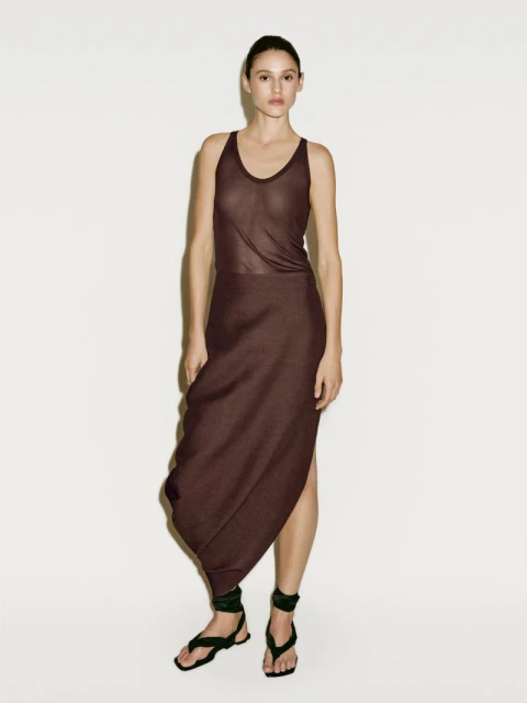 A woman in a dark brown tank top and a skirt with a draped hem stands against a white wall in a full shot, posing for fashion.
