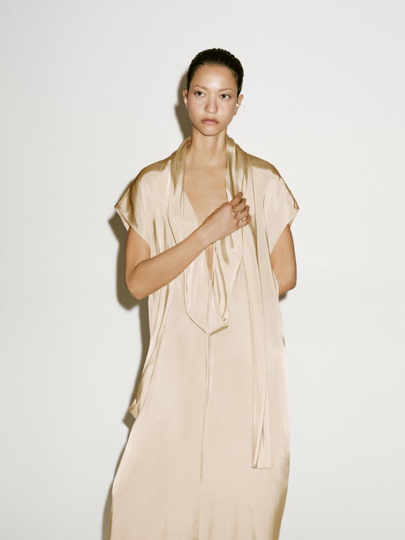Adult female posing indoors, wearing a light beige dress with a tie-neck, stands against a neutral background in a fashion studio setting.