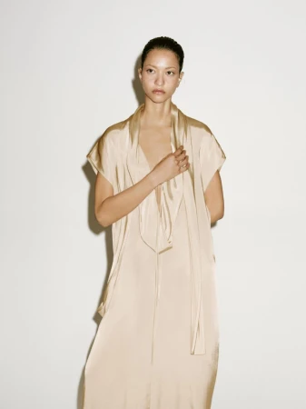 Adult female posing indoors, wearing a light beige dress with a tie-neck, stands against a neutral background in a fashion studio setting.