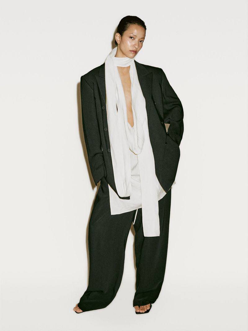 A woman in an oversized black blazer and pants, accessorized with a long white scarf, strikes a pose against a clean white backdrop.