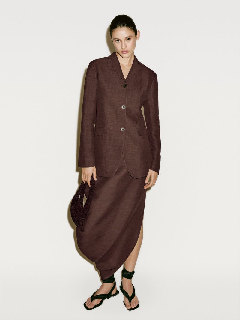 Elegant brunette woman in a brown linen suit stands against a neutral backdrop holding a small beaded bag, stylish sandals on her feet.