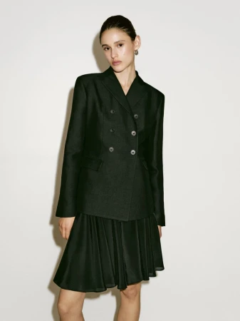 Model wearing a black double-breasted tailored blazer and pleated skirt against a plain studio backdrop.