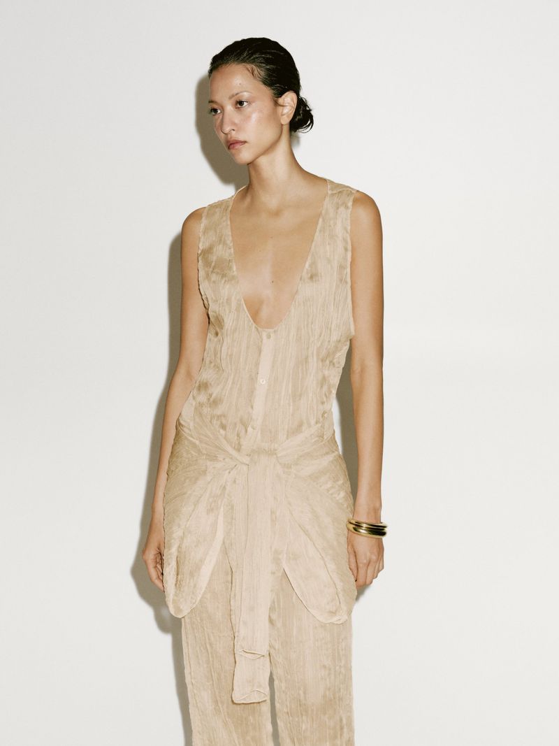 Elegant woman poses in a flowing, wrinkled jumpsuit against a white backdrop, showcasing neutral-toned fashion and subtle studio lighting.