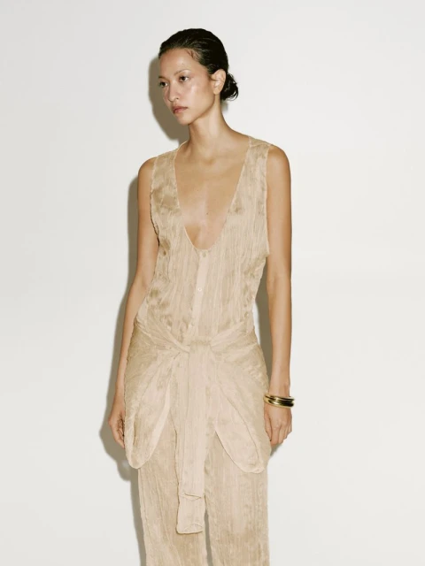 Elegant woman poses in a flowing, wrinkled jumpsuit against a white backdrop, showcasing neutral-toned fashion and subtle studio lighting.