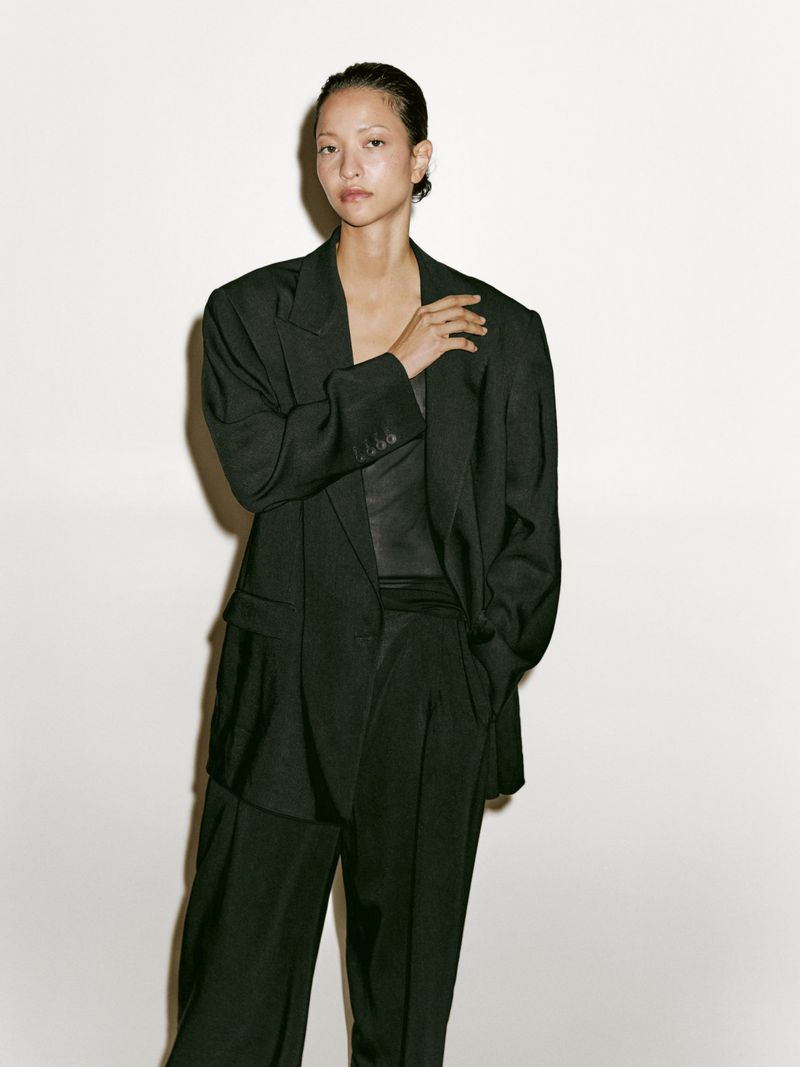 Woman in a dark green suit stands indoors against a white wall; modern, confident fashion.
