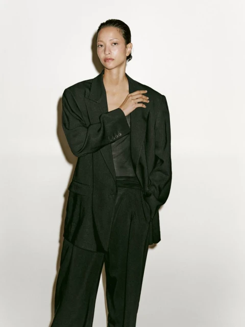 Woman in a dark green suit stands indoors against a white wall; modern, confident fashion.