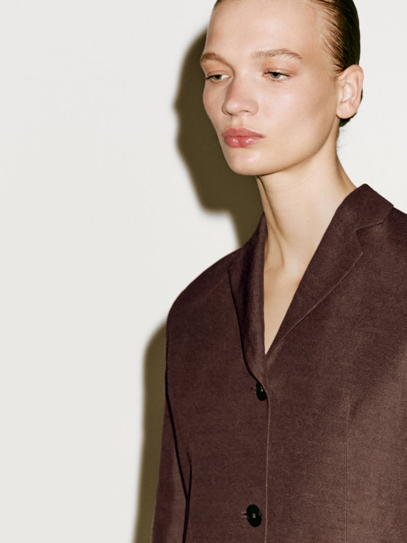 Close-up of a woman wearing a brown jacket against a white background. The jacket has black buttons and a V-neck collar.
