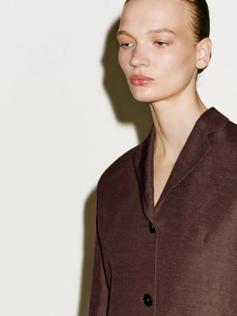 Close-up of a woman wearing a brown jacket against a white background. The jacket has black buttons and a V-neck collar.