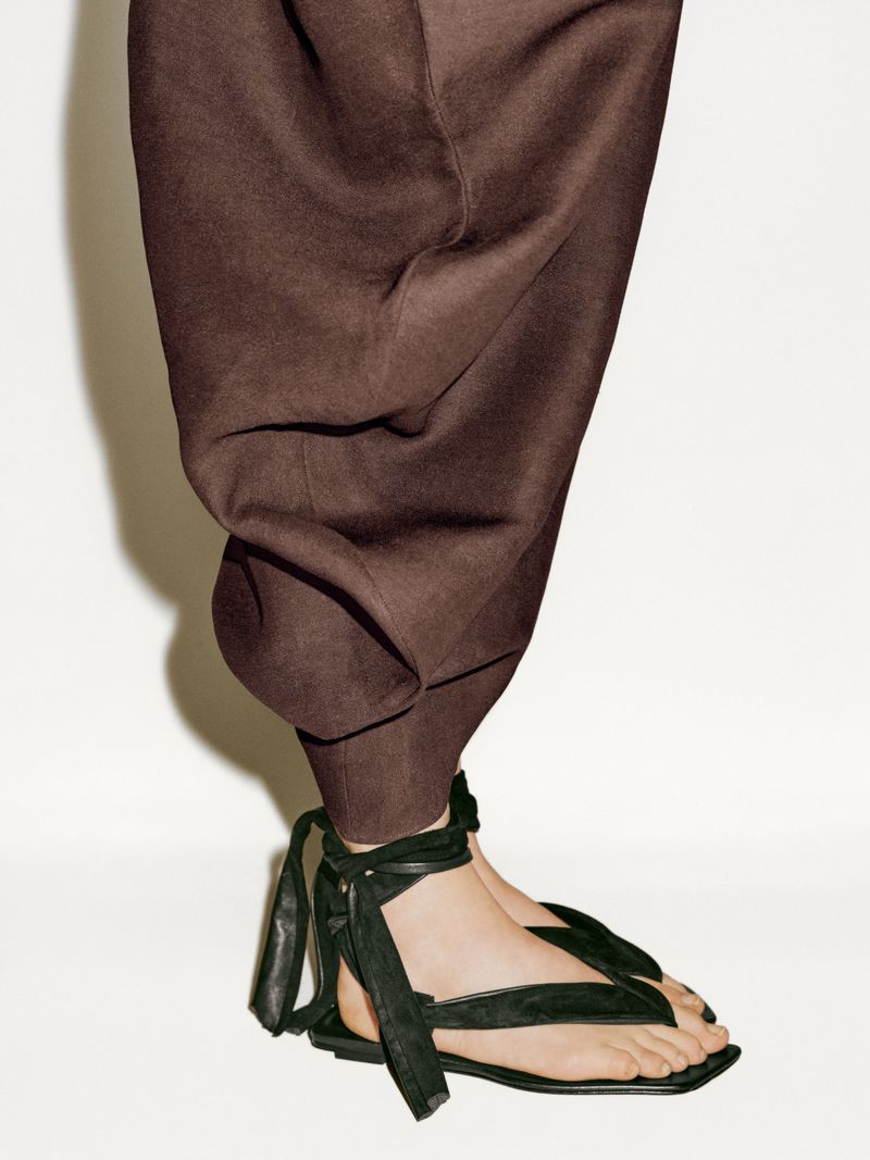 Lower portion of a woman wearing brown pants and black strappy sandals standing against a white background in a studio setting.