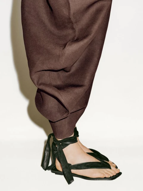 Lower portion of a woman wearing brown pants and black strappy sandals standing against a white background in a studio setting.