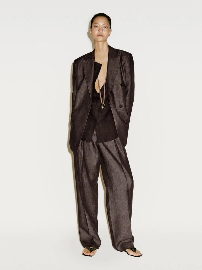 Model wearing a dark oversized blazer and matching trousers with a long pendant necklace against a plain white background.