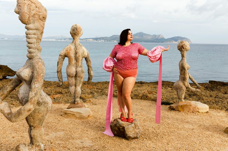 Woman in a pink outfit poses in front of sculptures and ocean on a rocky beach on a cloudy day, creating a surreal fashion image.