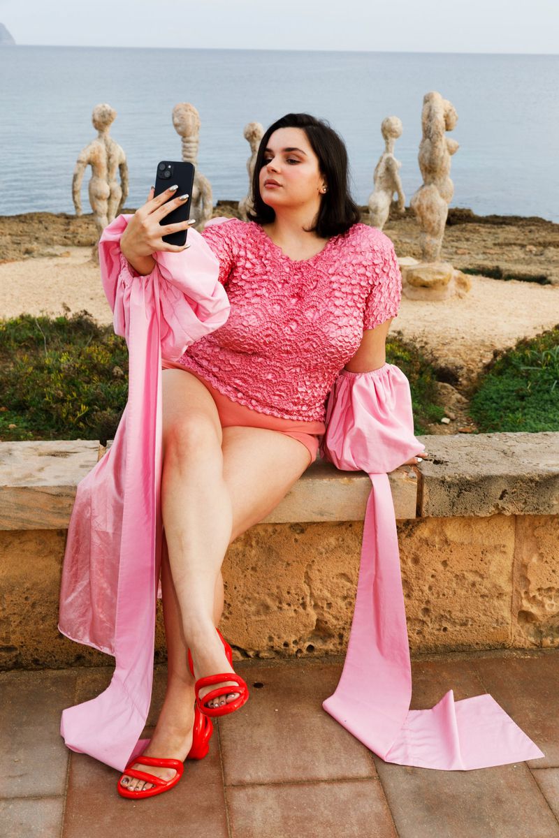 A woman in a pink outfit takes a selfie outdoors near stone sculptures and the ocean, capturing a vibrant and modern scene.