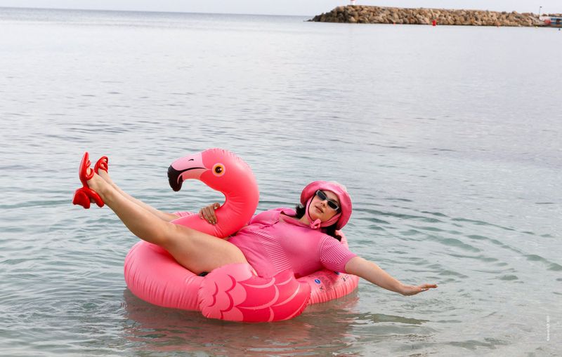 Woman in pink outfit floats on a flamingo in the ocean with a rocky breakwater in the background on a bright, sunny day.