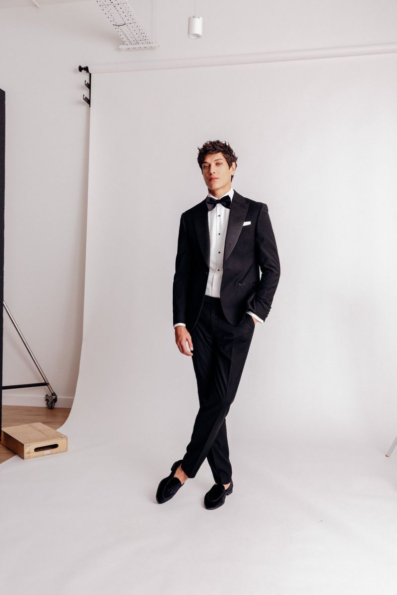 Elegant man in black tuxedo and bow tie stands in a photo studio against a white backdrop, casting a sophisticated figure in formal wear.