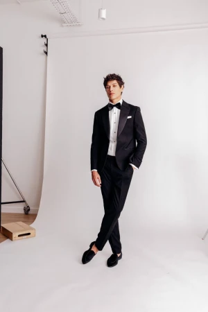 Elegant man in black tuxedo and bow tie stands in a photo studio against a white backdrop, casting a sophisticated figure in formal wear.