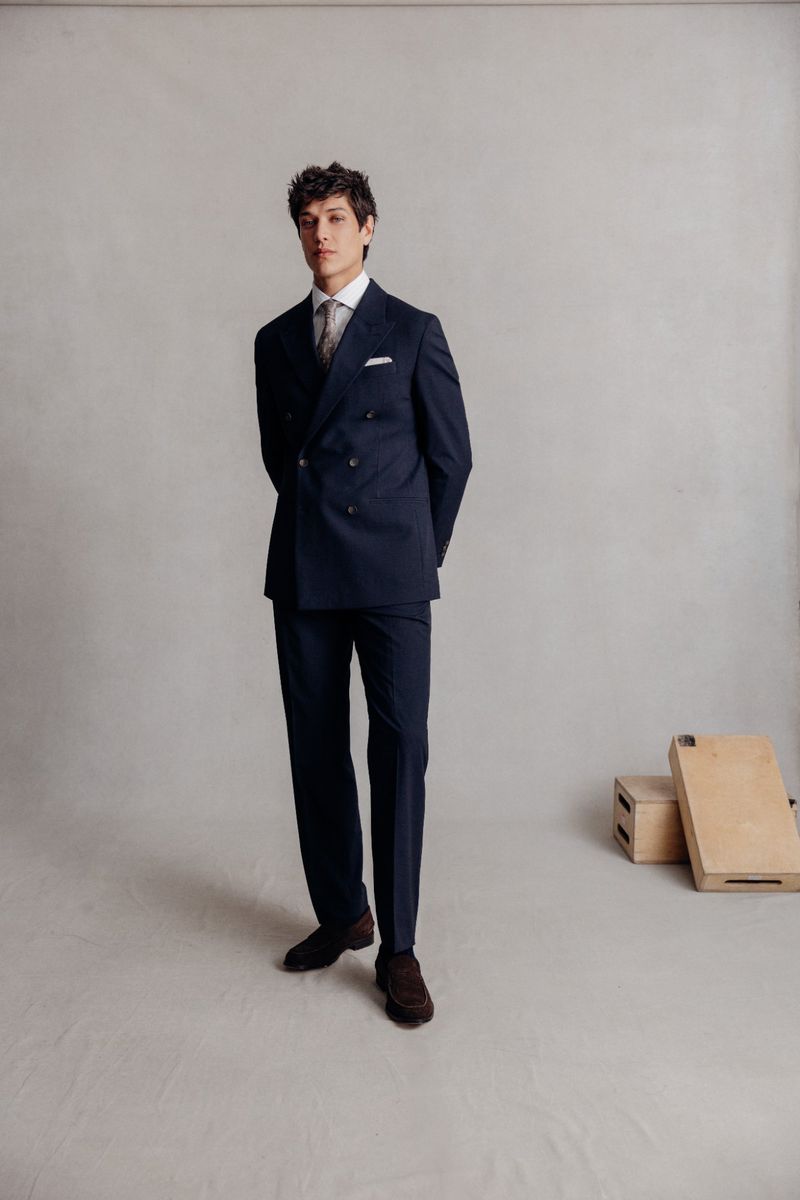 A young adult male model poses in a double-breasted navy suit, light shirt, and patterned tie against a light gray studio backdrop.