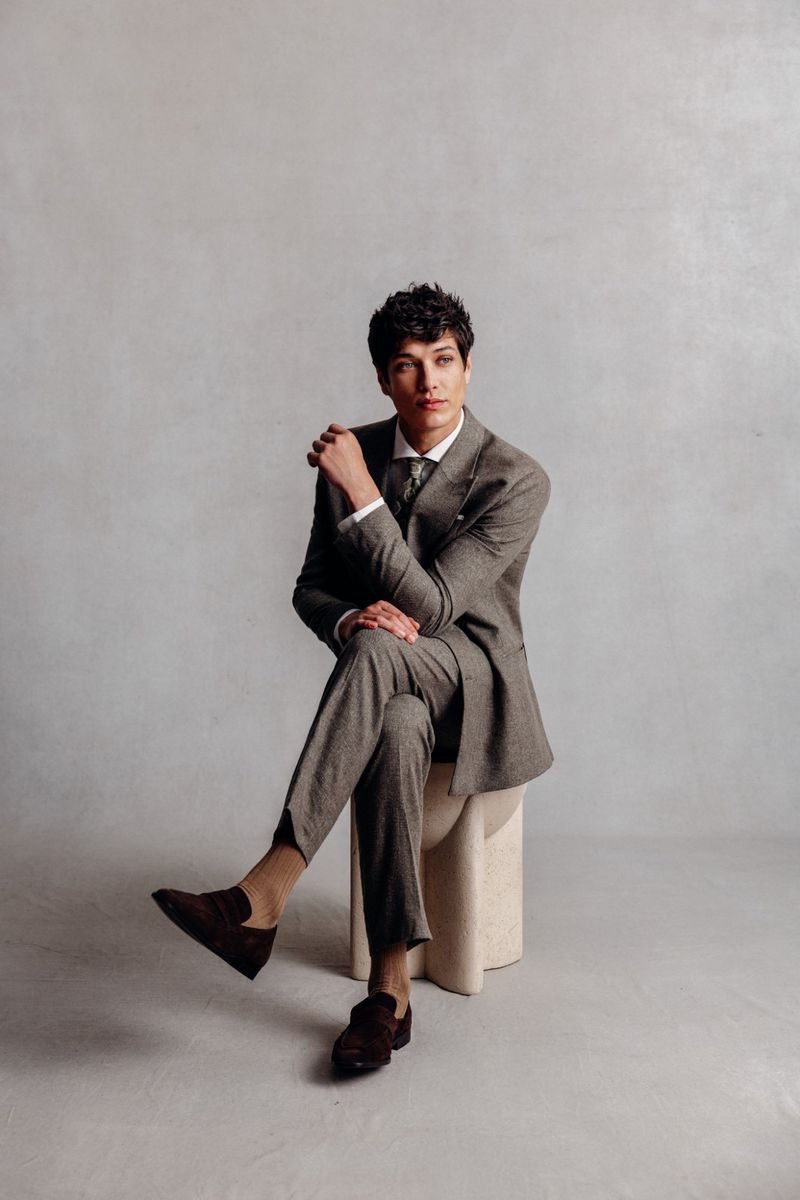 Young man in a gray suit sits on a light-beige stool in a studio. He is wearing brown suede loafers and has his legs crossed.