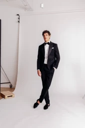 Full-length portrait of a male model wearing a black tuxedo and bow tie standing against a white studio background.