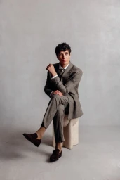 Aaron Gatward wearing a tailored taupe suit seated on a low stone stool against a textured grey studio backdrop.