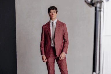 Male model in a burgundy pinstripe suit and tie standing against a light grey studio background.