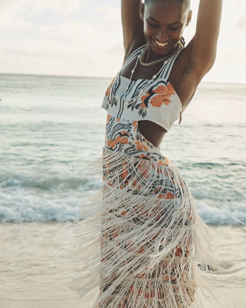 Woman in floral print dress with fringe poses on beach with ocean backdrop. Natural light captures the scene with a happy expression.