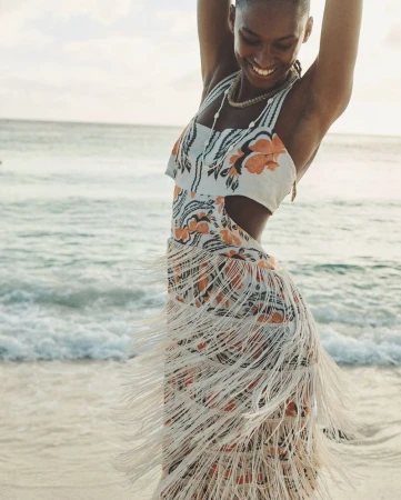 Woman in floral print dress with fringe poses on beach with ocean backdrop. Natural light captures the scene with a happy expression.