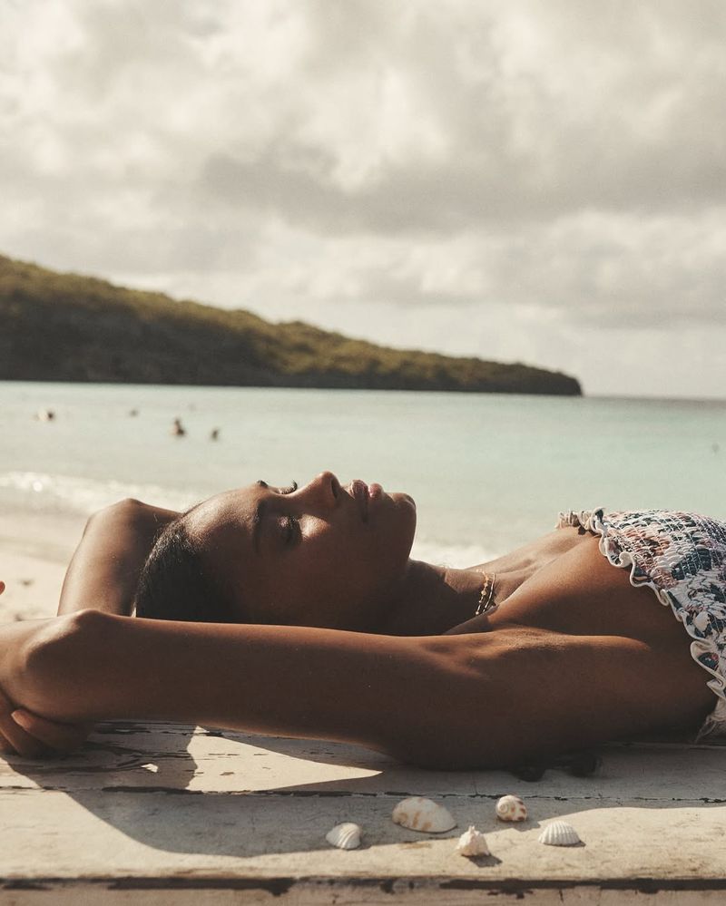 Woman in swimwear lounges on a wooden surface with seashells on a beach near the ocean under a partly cloudy sky.