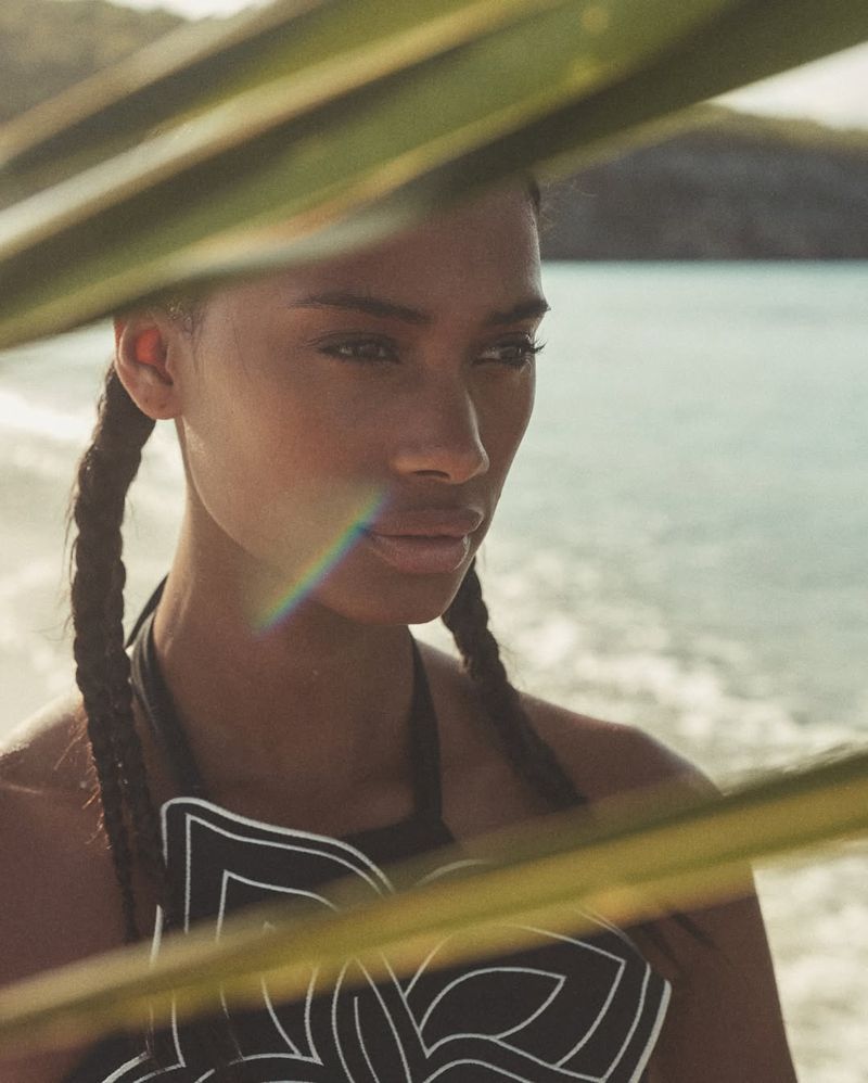 Close-up of a dark-skinned woman with braided pigtails and a halter top, gazing pensively near a beach with partial palm frond framing.