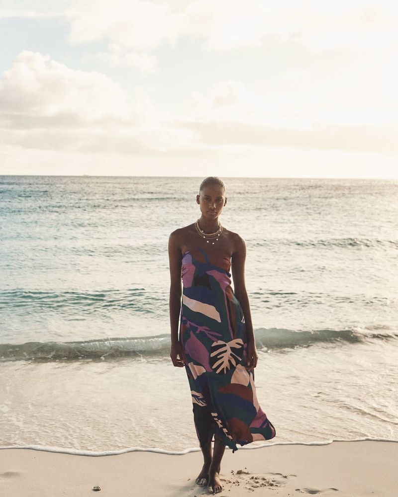A solitary Black woman poses on a sandy beach in a vibrant, patterned sundress, with the ocean and sky creating a serene backdrop.