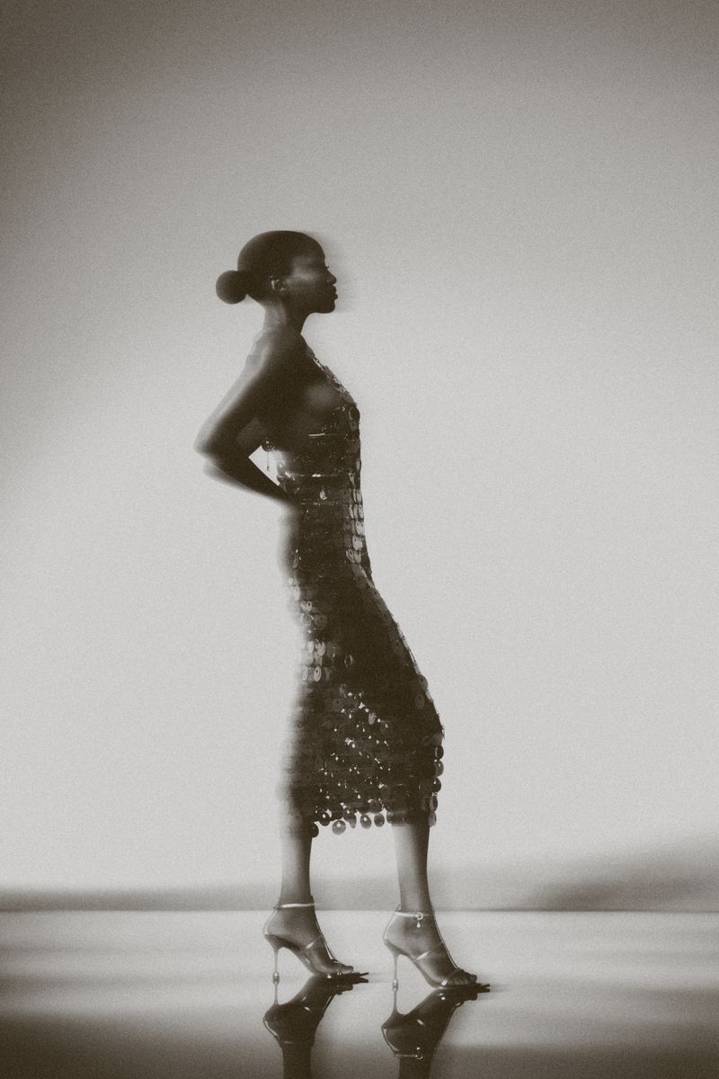 Monochrome portrait of a Black woman in a shimmering dress and high heels, striking a pose against a blurred background in a studio setting.