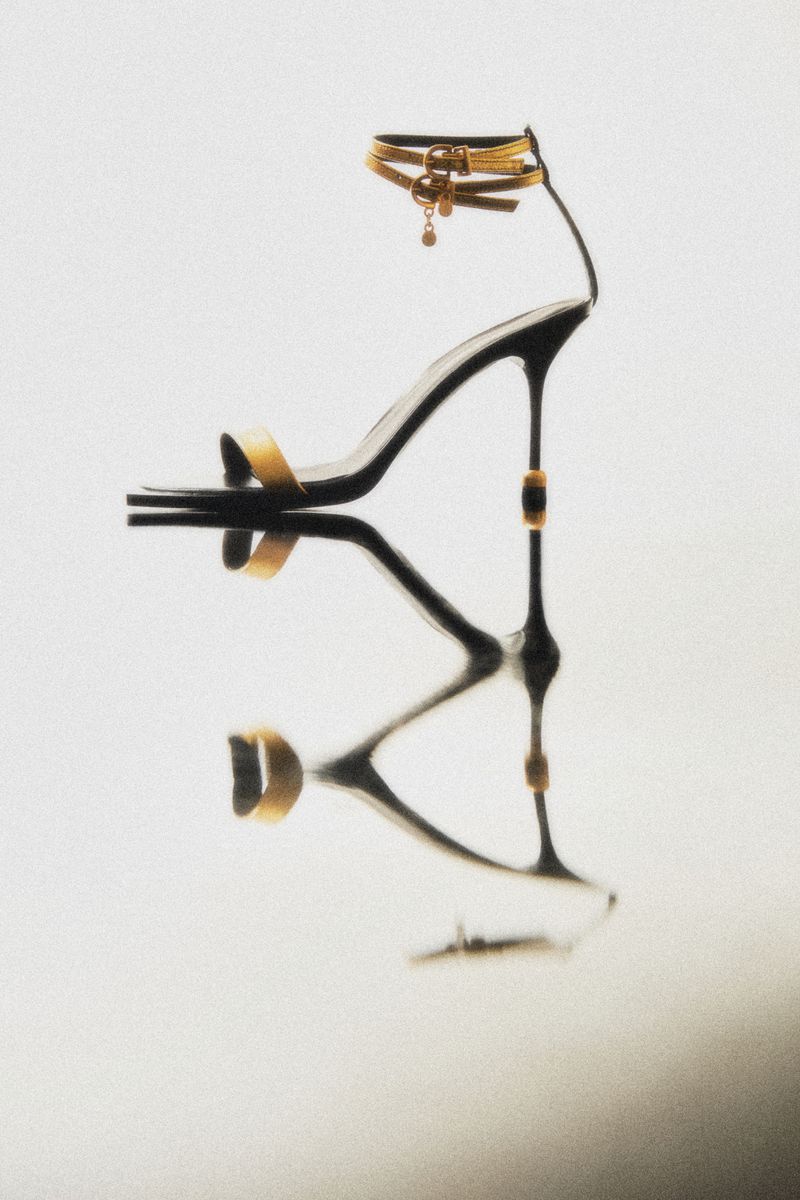 Elegant black stiletto heel sandal with gold accents and ankle straps on a reflective surface, studio lit, fashion footwear.