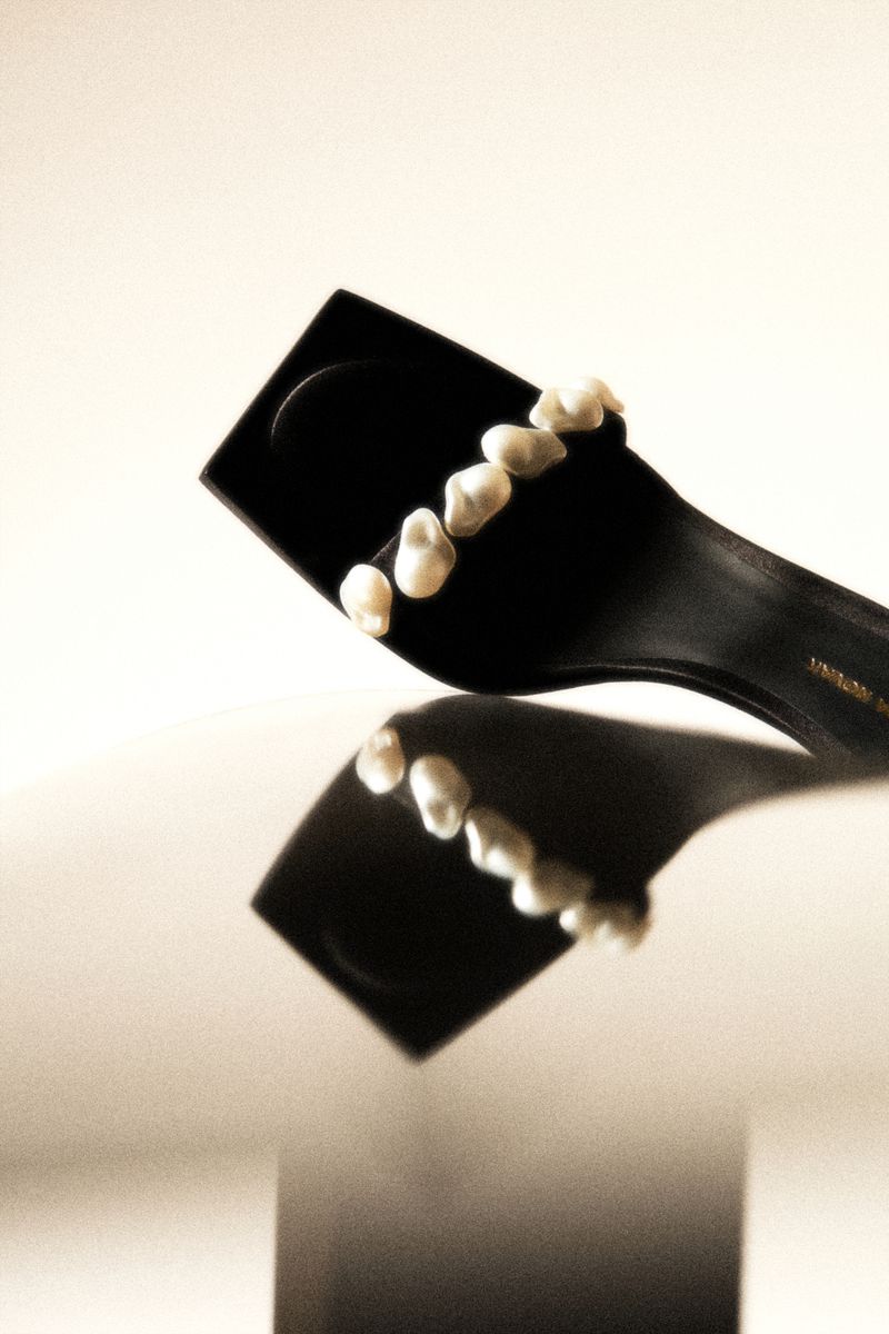 Elegant black high heel sandal embellished with pearls, reflected on a glossy surface against a soft, neutral background.