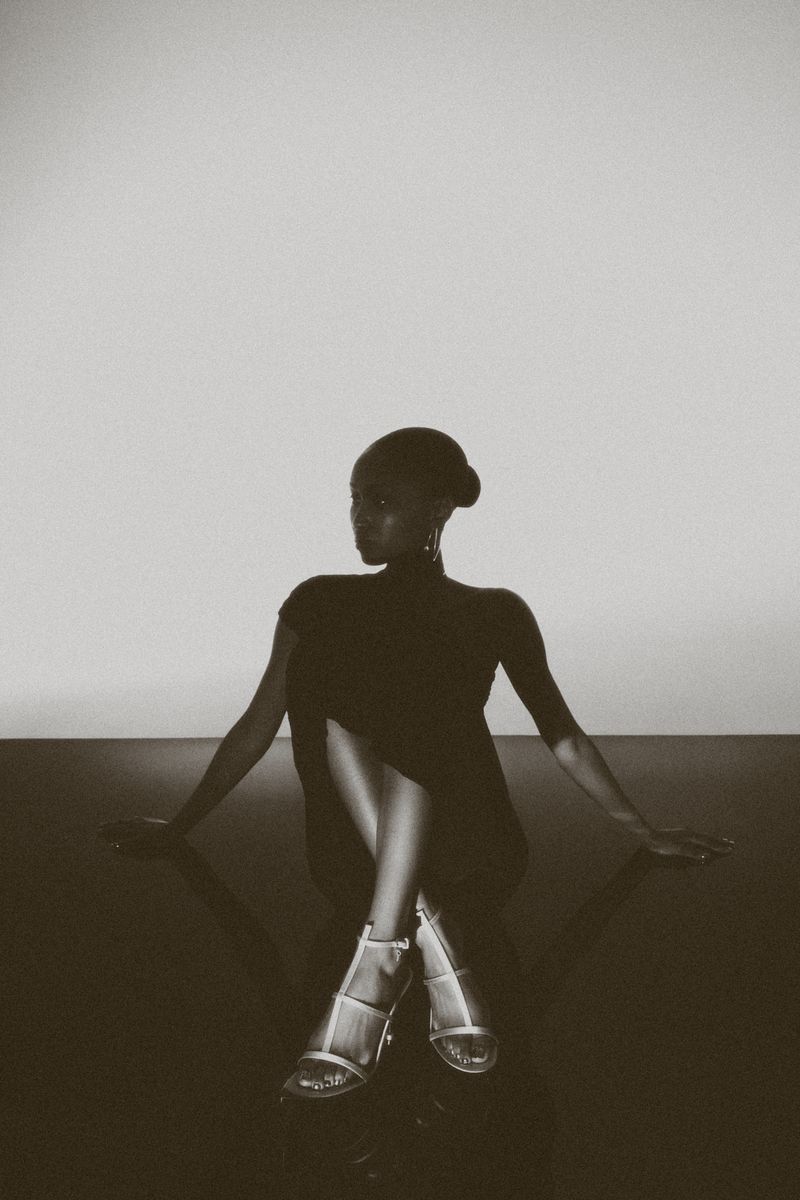 Elegant dark-skinned woman sits in silver heels and a short black dress against a gray backdrop, exuding minimalist style.