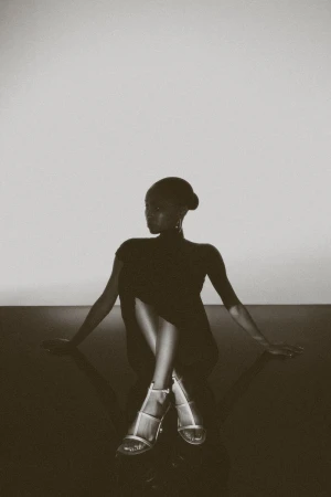 Elegant dark-skinned woman sits in silver heels and a short black dress against a gray backdrop, exuding minimalist style.
