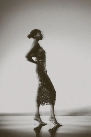 Model posing in profile wearing a textured disc midi dress and stiletto heels against a luminous studio backdrop.
