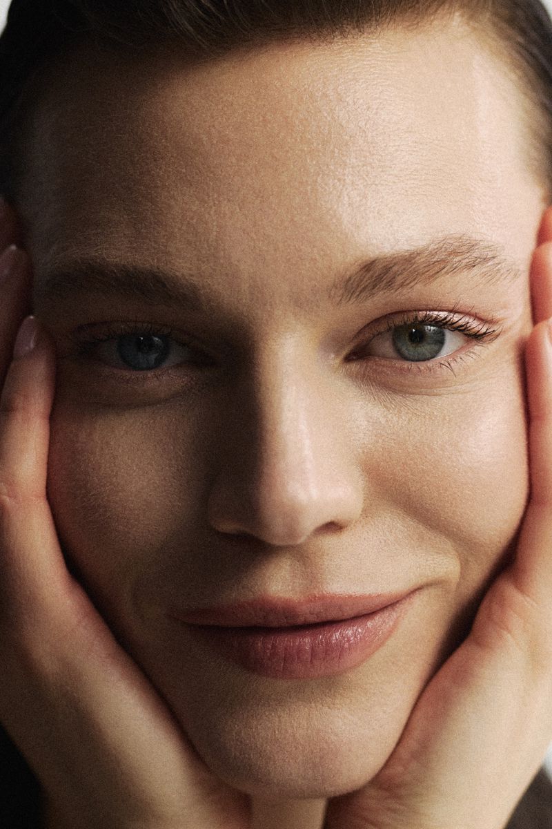 Close-up portrait of a fair-skinned adult woman with hands framing her face, featuring cool-toned skin, and a neutral facial expression.